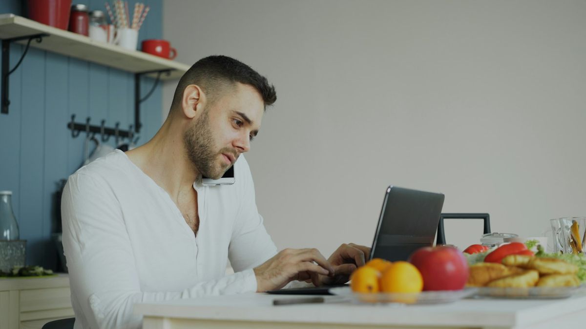 Hombre al teléfono mientras revisa sus cuentas en su laptop, en la cocina